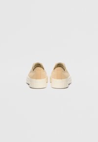 Beiga canvas loafers med vit gummisula och texturerad baksidedetalj, sedda från baksidan, som visar en minimalistisk design.