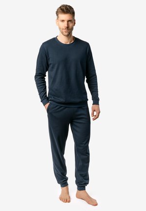 Man standing barefoot wearing a dark blue long-sleeve sweatshirt and matching jogger pants on a white background.