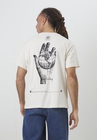 Bežs kokvilnas T-krekls ar melnu grafiku, kurā attēlota roka, kas tur globusu uz muguras, ar tekstu un cipariem zem tā. Īsas piedurknes, brīvs piegriezums.
