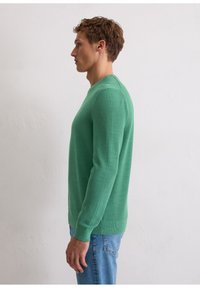 Grüner Strickpullover mit rundem Ausschnitt, strukturierter Stoff, langen Ärmeln und gerippten Bündchen, kombiniert mit hellblauen Jeans.