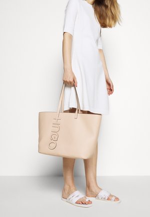Beige leather tote bag with embossed logo, square shape, two long handles, paired with a white dress and white strappy sandals.