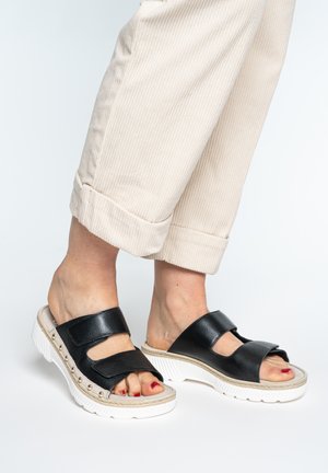 Feet wearing black open-toe slide sandals with two straps and white textured soles, paired with beige cropped pants.