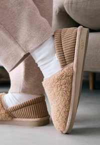 Braune Plüschschuhe mit gerippten Stoffakzenten, weichem Textil und einer beigen Gummisohle, getragen mit weißen Socken. Der Markenname "NEXT" ist sichtbar.