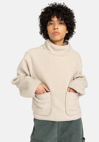 Persona con cabello rizado que lleva un pullover de forro polar beige con cuello alto y grandes bolsillos frontales, con las manos dentro de los bolsillos, de pie contra un fondo blanco.