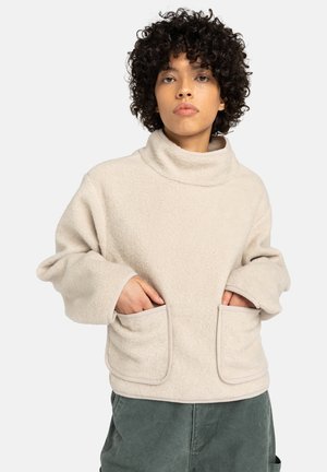 Persoon met krullend haar, gekleed in een beige fleece pullover met een hoge hals en grote voorzakken, met handen in de zakken, staand tegen een witte achtergrond.