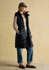 Sieviete, ģērbusies garā melnā izšūpu vestē, gaišā linsmeita garajās piedurknēs, sarullētās zilo džinsu biksēs, brūnās mokasīnās un rūtainajā cepurē, stāv ar rokām kabatās.