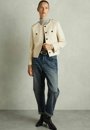 Romige cropped jas met zwarte knopen, twee zakken. Gecombineerd met blauwe jeans, opgerolde manchetten en zwarte enkellaarsjes met hak.