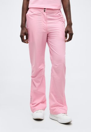 Nedre del af person iført løse pink bukser og hvide sneakers, stående mod en ensfarvet lys baggrund.