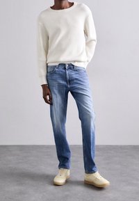 Vit ribbad tröja, ljusblå blekta jeans och beige sneakers. Jeans har rak passform; tröjan har en rundad hals.