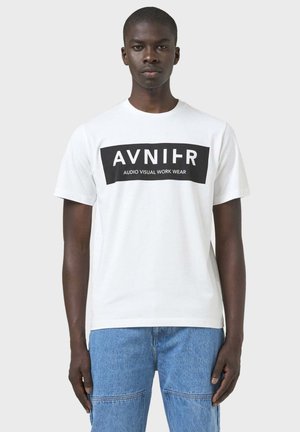 Mann trägt weißes T-Shirt mit schwarzem rechteckigem Logo mit der Aufschrift „AVNIIR AUDIO VISUAL WORK WEAR“ und blaue Jeans, steht vor grauem Hintergrund.