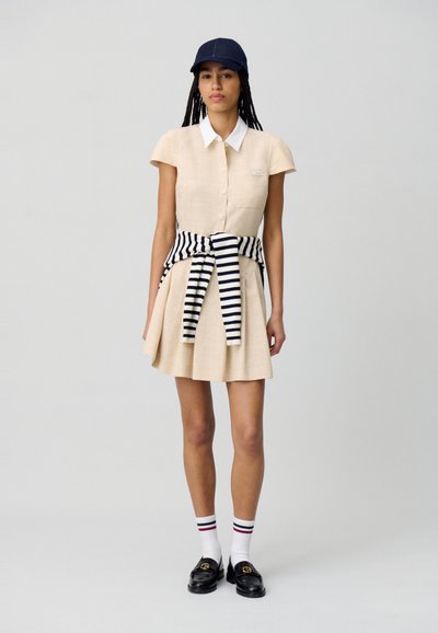 Robe beige à col avec des manches courtes, jupe plissée et cravate rayée noir et blanc. Associée à des chaussettes blanches et des mocassins noirs. Casquette marine.