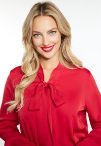 Rød silkebluse med indpakningsdesign og halsbånd, lange ærmer og en glat tekstur, båret med neutral makeup og stylet hår.
