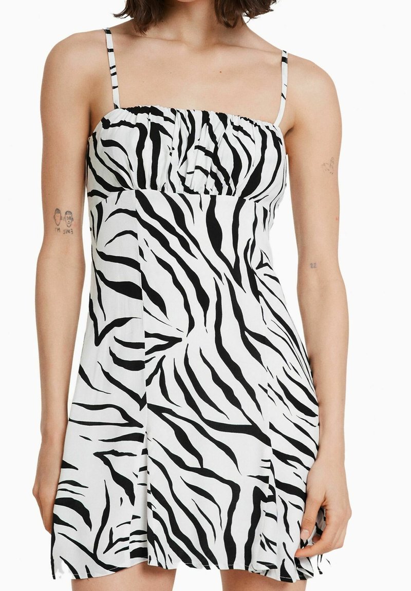 Femme portant une robe sans manches imprimé zèbre noir et blanc avec des fines bretelles, des tatouages visibles aux bras et les cheveux lâchés jusqu'aux épaules.