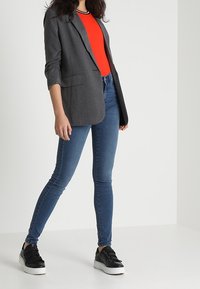 Person standing wearing a gray blazer, red top, blue skinny jeans, and black sneakers with white soles against a plain background.