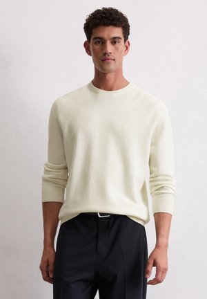 Mann mit kurzem, lockigem Haar, der einen cremefarbenen Strickpullover und schwarze Hosen trägt, steht vor einem schlichten weißen Hintergrund.