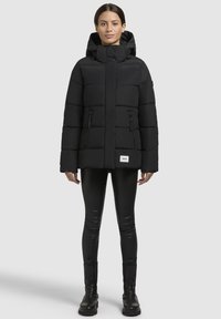 Veste noire matelassée avec capuche, dotée de poches zippées, d'un col montant et d'un patch logo en bas. Portée par-dessus des leggings noirs et des bottes.