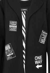 Schwarze Jacke mit weißem Text und grafischen Aufnähern. Verfügt über ein gestreiftes Futter, einen Reverskragen und verschiedene Drucklabels auf der Vorderseite.