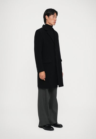 Cappotto di lana nero con colletto a punta, chiusura con un bottone singolo, tasche laterali, abbinato a pantaloni grigi larghi e scarpe di pelle nere.