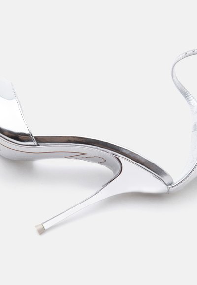 Ασημένιο σανδάλι με ψηλό τακούνι και λεπτό λουράκι στον αστράγαλο, εμφανιζόμενο σε λευκό φόντο.