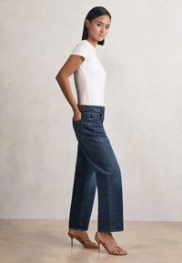 Kvinde stående sidelæns iført en hvid kortærmet skjorte, mørkeblå jeans med vide ben og brune høje hæle med remme mod en ensfarvet baggrund.