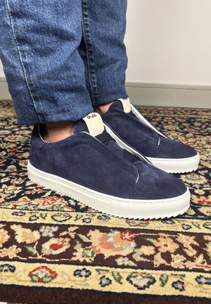 Person wearing navy blue suede slip-on sneakers with white soles and beige tongue tags, standing on a patterned carpet.