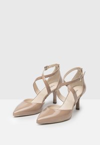 Beige patent leather heels with pointed toes and a 5 cm block heel. Features crisscross ankle straps and a smooth, glossy finish.