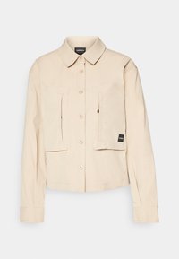 Giacca beige con bottoni e colletto, due tasche anteriori a toppa e una piccola etichetta nera sul lato sinistro in basso. Tessuto dalla trama liscia.