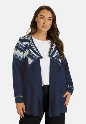 Femme portant un cardigan bleu marine avec des motifs géométriques blancs et bleu clair sur une chemise blanche et un pantalon noir.