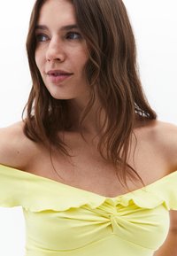 Top off-the-shoulder di un brillante giallo con colletto a ruffles, texture morbida e dettaglio arricciato al centro.