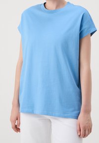 Ljusblå bomullströja med rund hals och korta ärmar. Något lös passform, slät textur och rak fåll. Bärs med vita byxor.