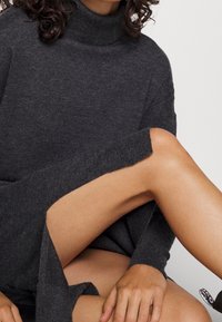 Ciemnoszary sweter z golfem o fakturowanym splotcie, w luźnym kroju z rozporkiem po bokach u dołu, odsłaniającym skórę.