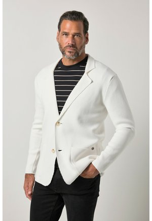 Man met kort donker haar en baard draagt een witte blazer over een zwart-wit gestreept shirt, met één hand in de zak, neutrale achtergrond.