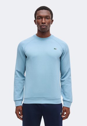 Homme portant un sweat-shirt bleu clair à manches longues et un pantalon bleu marine, debout devant un fond uni gris clair, face à la caméra.