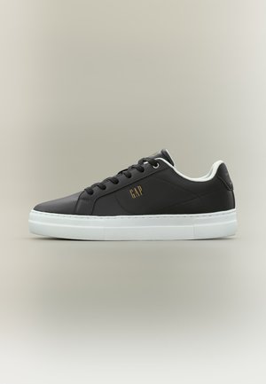 Scarpa da ginnastica nera bassa con suola bianca, lacci neri e logo "GAP" dorato sul lato, mostrata di profilo su sfondo neutro.