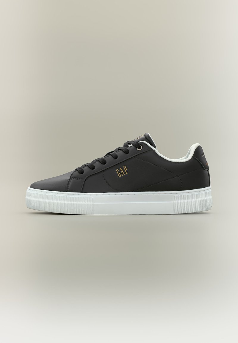 Baskets noires basses avec semelle blanche, lacets noirs et logo "GAP" doré sur le côté, présentées de profil sur un fond neutre.