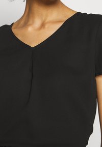 Top nero a maniche corte con scollatura a V, realizzato in tessuto morbido, con un delicato dettaglio a pieghe al centro e una texture liscia.