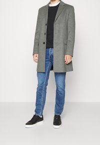 Cappotto di lana grigio con una vestibilità su misura, tre bottoni e rever a punta, abbinato a jeans scuri e sneakers nere.