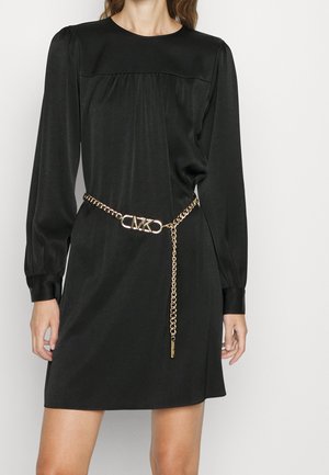 Kobieta nosząca czarną mini sukienkę z długim rękawem, bufkami na ramionach oraz złoty łańcuszkowy pasek z logo CMK w talii.