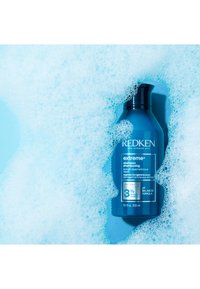 Blå flaska med Redken Extreme schampo, med en svart kork, märkt med produktinformation och omgiven av skummiga bubblor på en blå bakgrund.