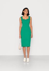 Vestido midi verde ajustado com decote quadrado, alças largas e detalhes de costura; combinado com sapatos brancos de deslizar e uma pequena mala bege riscada.