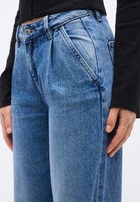 Κοντινό πλάνο ενός ατόμου που φορά ψηλόμεσα μπλε τζιν και μαύρο μακρυμάνικο τοπ, με τα χέρια χαλαρά δίπλα στο σώμα.