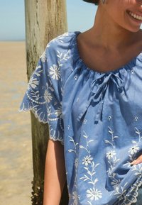 Kvinde iført en blå bluse med hvid blomsterbroderi og bølgede ærmer, stående ved en træpind på en sandstrand.