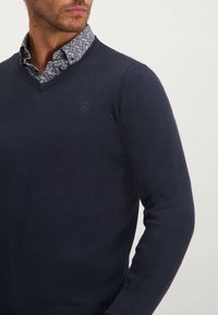 Marinblå v-ringad tröja gjord av mjukt stickat tyg, med ribbade detaljer på axlarna och en diskret logotyp på bröstet. Mönstrad skjorta synlig under.