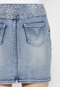 Jupe en denim avec un délavage bleu clair, dotée d'un design floral brodé au niveau de la taille, de deux poches arrière et d'une coupe droite.