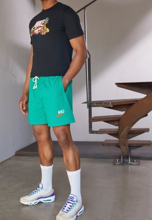 Camiseta negra con diseño de frutas, pantalones cortos verdes con textura y bolsillos, calcetines blancos y zapatillas beige con detalles morados, posicionados junto a una escalera.