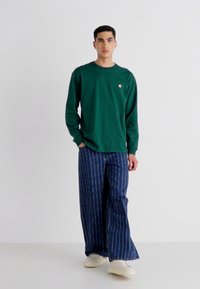 Green long-sleeve cotton T-shirt with a small logo on the chest, paired with wide-leg blue striped denim pants and white sneakers.