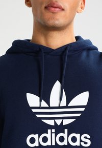 Man bär en marinblå hoodie med vit Adidas trefoil-logotyp och snoddar, synlig från läpparna till bröstet mot en enkel bakgrund.