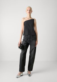 Top noir sans bretelles avec un design asymétrique, associé à un jean noir ample. Le mannequin tient un petit sac noir et porte des chaussures à talons blocs métalliques.