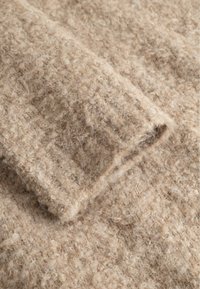 Artimos plano smėlio spalvos megztinio rankovė su tekstūruotu, kilpiniu vilnos audiniu ir ribinėta rankogaliu.