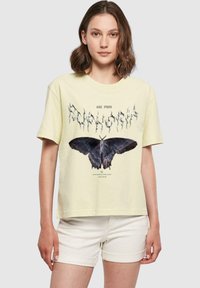 Gul bomullströja med en svart fjärilsgrafik och stiliserad text ovanför, kombinerad med ljusa shorts. Korta ärmar, avslappnad passform.